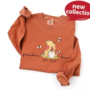 A long-sleeved orange t-shirt with a Winnie the Pooh graphic and text, along with a red speech bubble that says "new collection" and "EST. 2026".