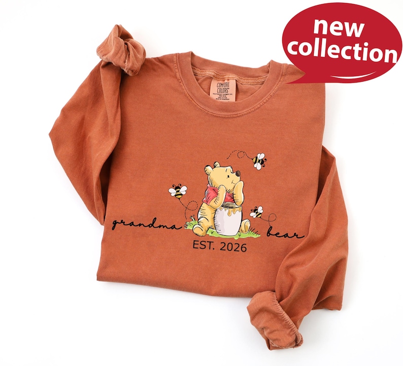 A long-sleeved orange t-shirt with a Winnie the Pooh graphic and text, along with a red speech bubble that says "new collection" and "EST. 2026".