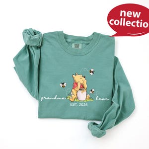 A green t-shirt with a Winnie the Pooh character and the text "Grandma Bear" printed on it, along with the text "EST. 2026" below the image.