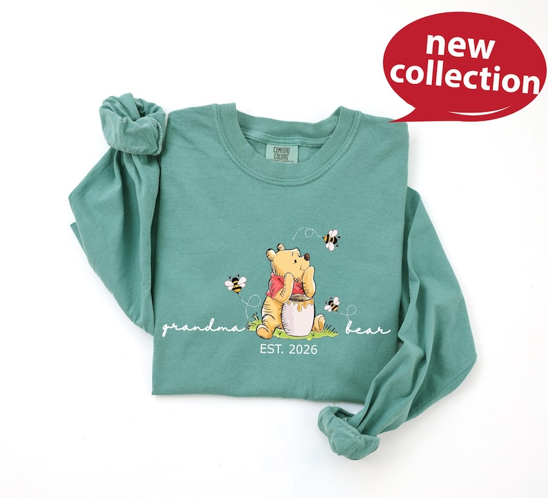 A green t-shirt with a Winnie the Pooh character and the text "Grandma Bear" printed on it, along with the text "EST. 2026" below the image.