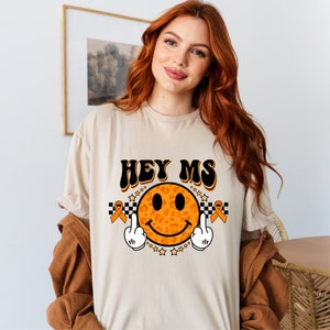 May include: A beige t-shirt with the text "HEY MS" in black and a leopard print smiley face with orange and black middle fingers. The smiley face is surrounded by orange awareness ribbons and black and white checkered pattern.