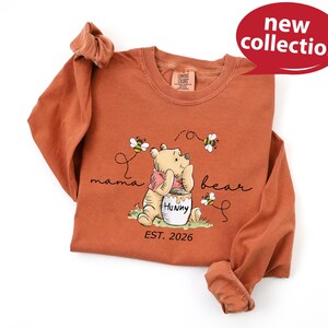 A red long-sleeved t-shirt with a Winnie the Pooh graphic and text, along with a red speech bubble that says "new collection".