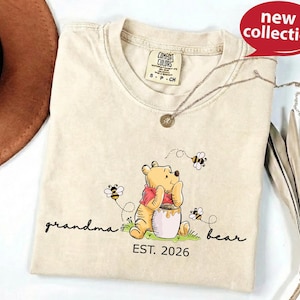A beige t-shirt with a Winnie the Pooh-themed design, including a bear, honey pot, and bees, along with the text "Grandma Bear" and "EST. 2026".