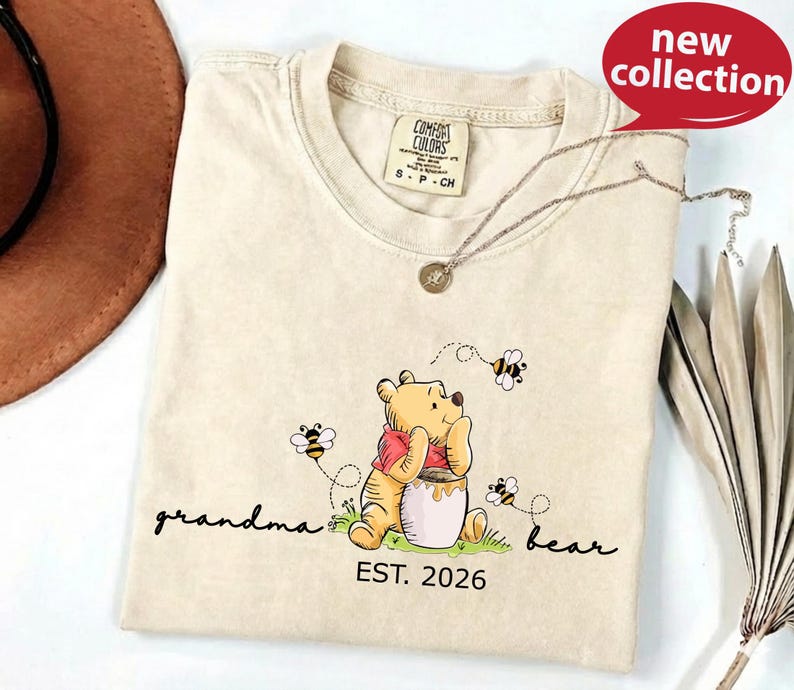 A beige t-shirt with a Winnie the Pooh-themed design, including a bear, honey pot, and bees, along with the text "Grandma Bear" and "EST. 2026".