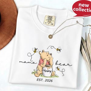 A white t-shirt with a Winnie the Pooh-themed design, including the character Winnie the Pooh and honey pot, along with the text "mama bear" and "HUNNY" printed on it. The t-shirt is displayed on a white surface, with a brown hat and a necklace visible in the background.
