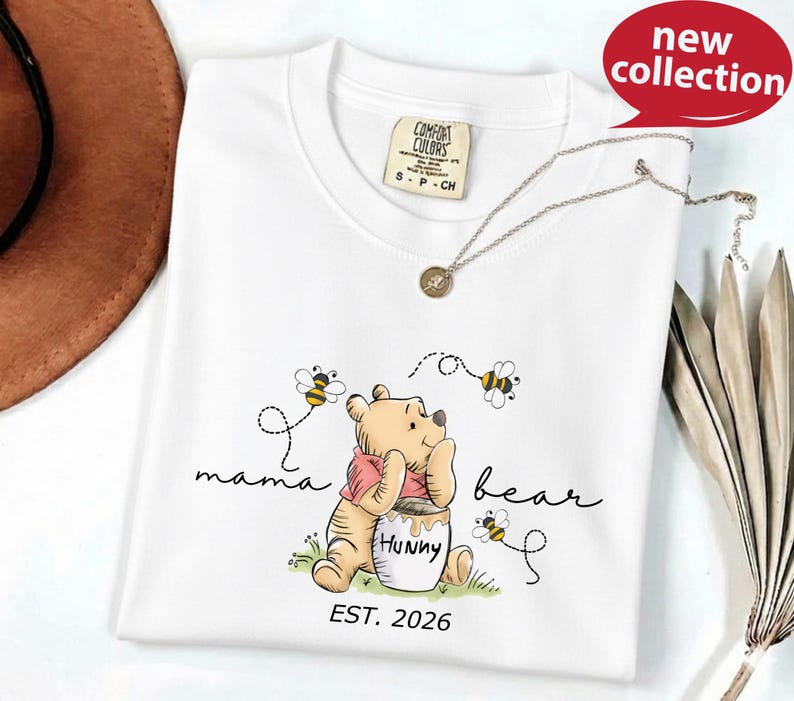 A white t-shirt with a Winnie the Pooh-themed design, including the character Winnie the Pooh and honey pot, along with the text "mama bear" and "HUNNY" printed on it. The t-shirt is displayed on a white surface, with a brown hat and a necklace visible in the background.