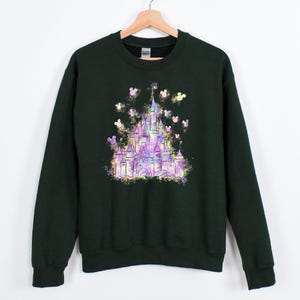 Puede incluir: Sudadera verde oscuro con un gr&aacute;fico de castillo colorido y siluetas de Mickey Mouse. El dise&ntilde;o del castillo es en tonos morado, rosa y amarillo. La sudadera tiene mangas largas y cuello redondo.