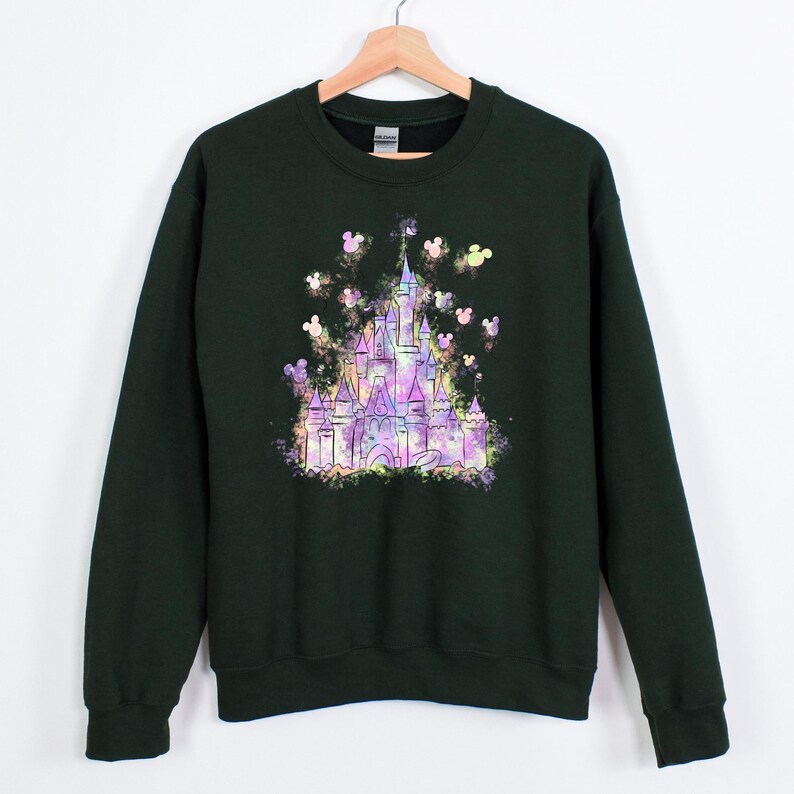 Puede incluir: Sudadera verde oscuro con un gr&aacute;fico de castillo colorido y siluetas de Mickey Mouse. El dise&ntilde;o del castillo es en tonos morado, rosa y amarillo. La sudadera tiene mangas largas y cuello redondo.