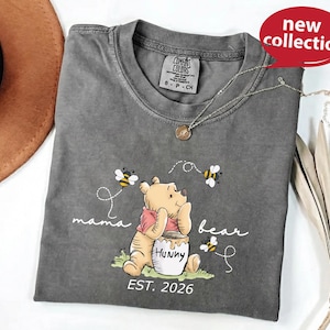 A gray t-shirt with a Winnie the Pooh character printed on it, along with the text "mama bear" and "humpty" printed below. The t-shirt is displayed on a white surface, with a brown hat and some leaves in the background.