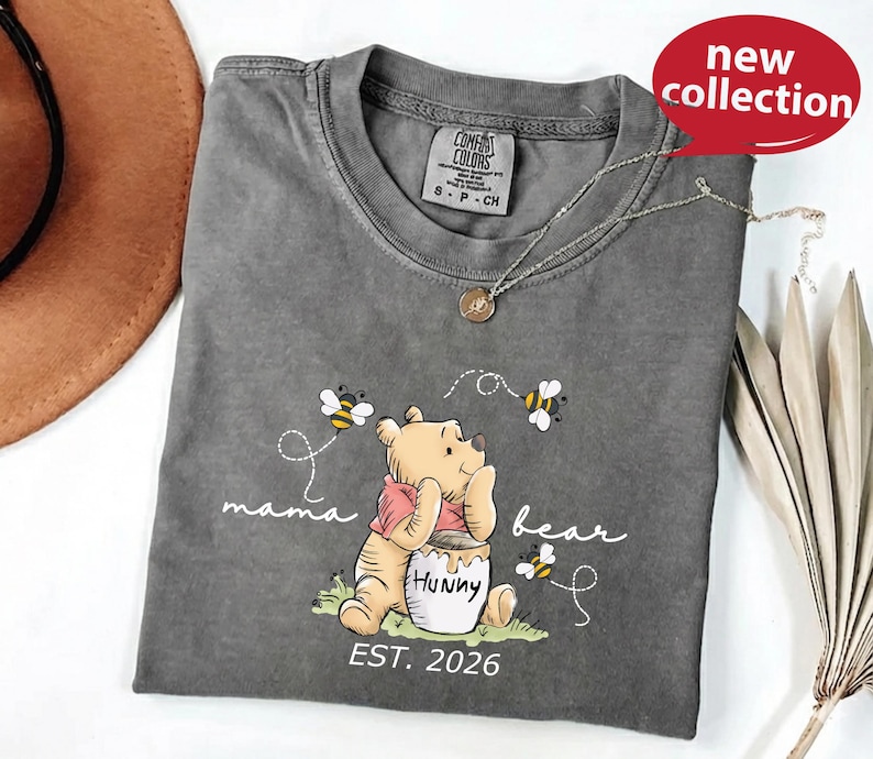 A gray t-shirt with a Winnie the Pooh character printed on it, along with the text "mama bear" and "humpty" printed below. The t-shirt is displayed on a white surface, with a brown hat and some leaves in the background.