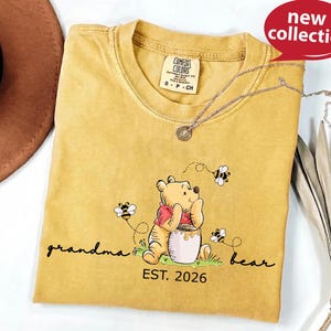 A yellow t-shirt with a Winnie the Pooh graphic and the text "Grandma Bear" printed on it, along with a necklace and a hat.