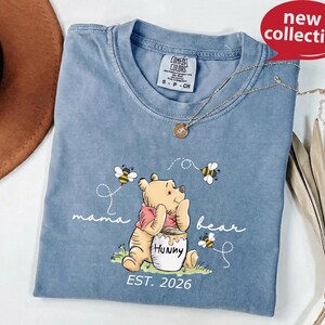 A blue t-shirt with a Winnie the Pooh graphic and the text "mama bear" printed on it, along with the text "EST. 2026" below the image. The t-shirt is displayed on a white surface, and there is a brown hat and some leaves in the background.
