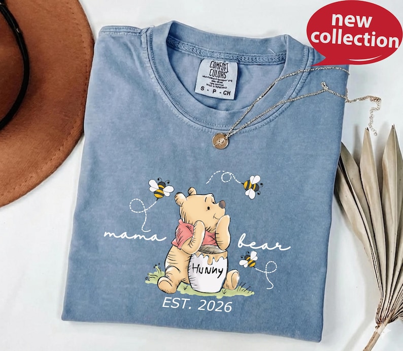 A blue t-shirt with a Winnie the Pooh graphic and the text "mama bear" printed on it, along with the text "EST. 2026" below the image. The t-shirt is displayed on a white surface, and there is a brown hat and some leaves in the background.