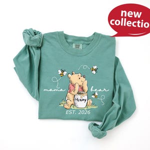 A green long-sleeved t-shirt with a Winnie the Pooh character and honey pot design, along with the text "mama bear" and "Est. 2026".