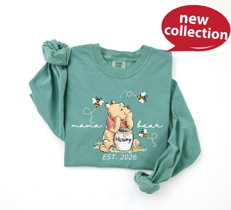 A green long-sleeved t-shirt with a Winnie the Pooh character and honey pot design, along with the text "mama bear" and "Est. 2026".