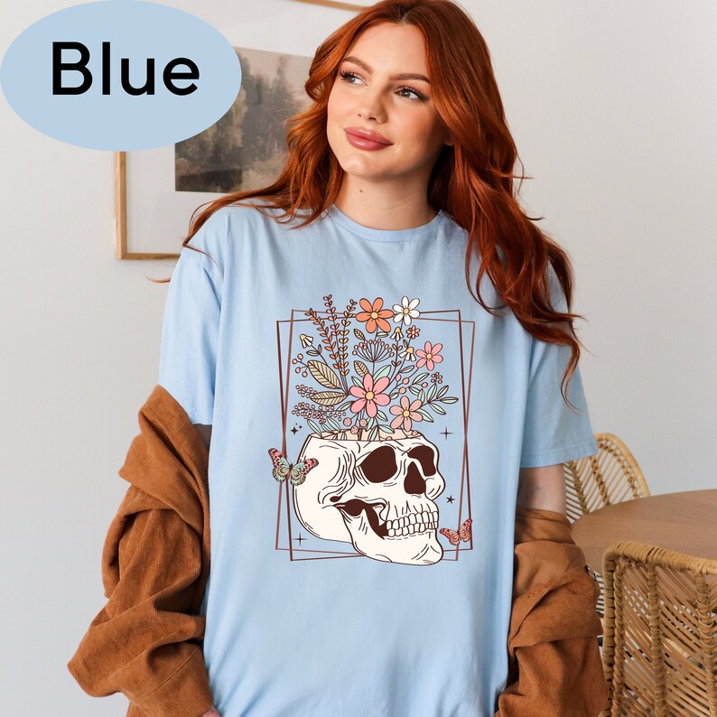 May include: A light blue t-shirt with a graphic of a skull with flowers growing out of the top. The graphic is framed in a gold rectangle with butterflies on either side.