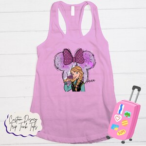 May include: A light pink tank top with a graphic of Anna from Frozen within a Mickey Mouse ear outline. The design includes a purple bow, snowflakes, and the name "Anna". A pink suitcase with travel stickers is in the image. Text on the bottom reads "Custom Disney Trip Tank Tops."