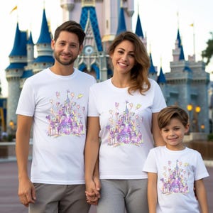 Puede incluir: Camisetas blancas con un dise&ntilde;o de castillo colorido. El dise&ntilde;o presenta un castillo con detalles en morado, rosa y amarillo, junto con globos en forma de coraz&oacute;n. Las camisetas son usadas por una familia frente a un castillo.