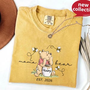 A yellow t-shirt with a Winnie the Pooh design and the text "mama bear" printed on it, along with a necklace and a hat.