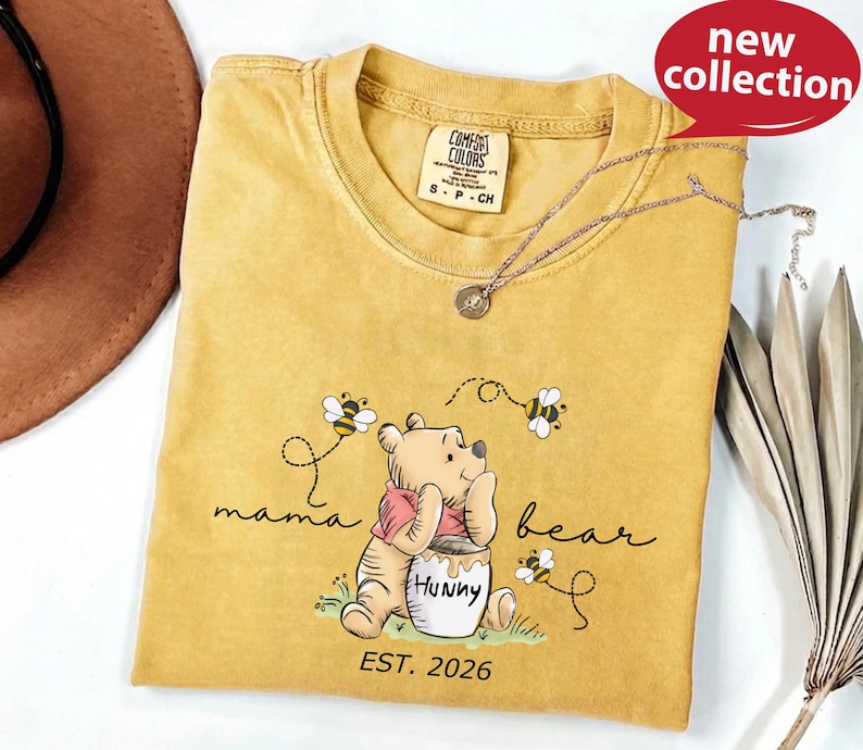 A yellow t-shirt with a Winnie the Pooh design and the text "mama bear" printed on it, along with a necklace and a hat.