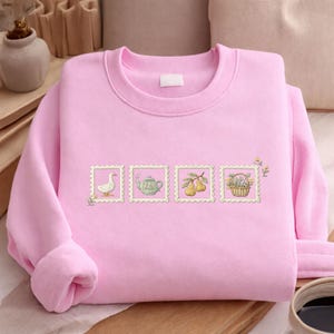 A pink sweater with a row of four small illustrations on it, including a duck, teapot, basket of fruit, and flowers.
