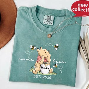A green t-shirt with a Winnie the Pooh design and the text "mama bear" printed on it, along with the text "EST. 2026" below the image.