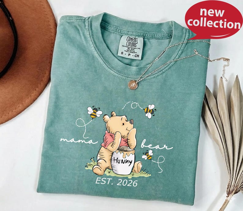 A green t-shirt with a Winnie the Pooh design and the text "mama bear" printed on it, along with the text "EST. 2026" below the image.