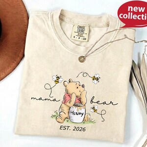 A beige t-shirt with a Winnie the Pooh graphic and the text "mama bear" printed on it, along with a necklace and a brown hat.