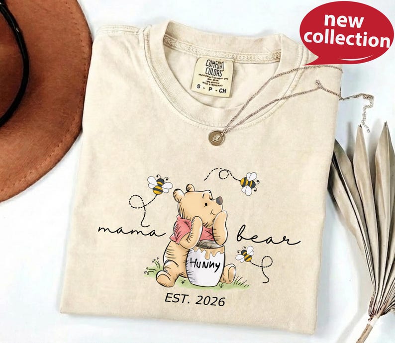 A beige t-shirt with a Winnie the Pooh graphic and the text "mama bear" printed on it, along with a necklace and a brown hat.