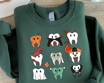May include: A dark green sweatshirt with a Halloween-themed design featuring various teeth dressed up in costumes. The teeth are wearing a pumpkin, a witch's hat, a mummy costume, a clown hat, a ghost costume, a vampire costume, and a skeleton costume. The sweatshirt has a crew neck and long sleeves.