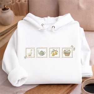 A white hoodie with a floral embroidered design on the front, featuring a teapot, a duck, and a basket of fruit.