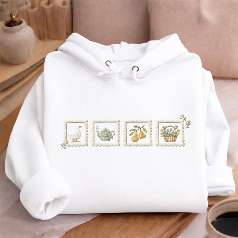 A white hoodie with a floral embroidered design on the front, featuring a teapot, a duck, and a basket of fruit.