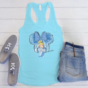 May include: A turquoise tank top with a Cinderella graphic. The graphic features a blue bow, Cinderella, and the word "Cinderella". Grey trainers and blue jeans are also in the image.