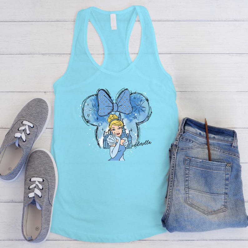 May include: A turquoise tank top with a Cinderella graphic. The graphic features a blue bow, Cinderella, and the word "Cinderella". Grey trainers and blue jeans are also in the image.