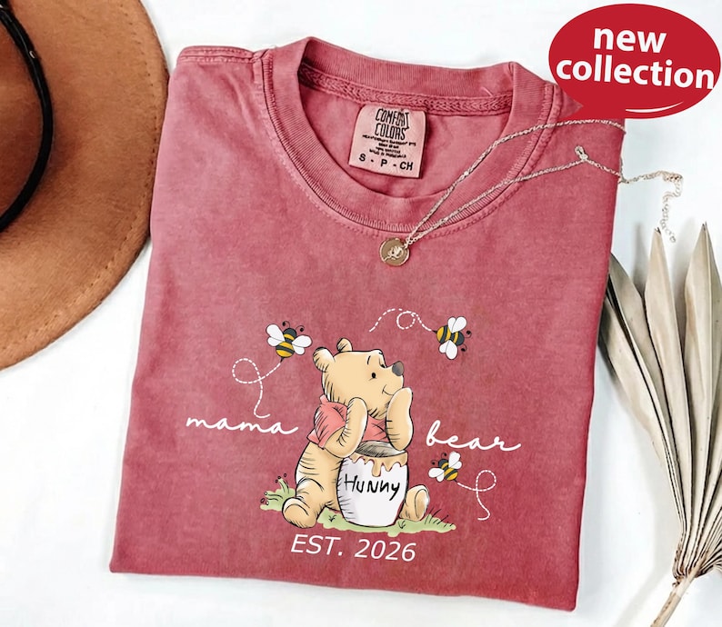 A pink t-shirt with a Winnie the Pooh character and the text "mama bear" printed on it, along with a necklace and a hat.