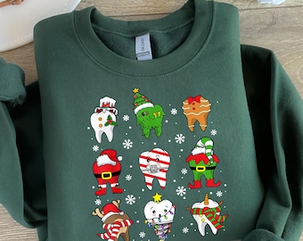 May include: A dark green sweatshirt featuring a festive design of cartoon teeth dressed in Christmas attire. The teeth are decorated as Santa, elves, gingerbread men, and more, with snowflakes scattered around.