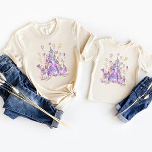 Puede incluir: Dos camisetas color crema con un dise&ntilde;o de castillo en acuarela en morado, rosa y azul. Las camisetas se combinan con vaqueros azules. El dise&ntilde;o del castillo incluye globos en forma de coraz&oacute;n.