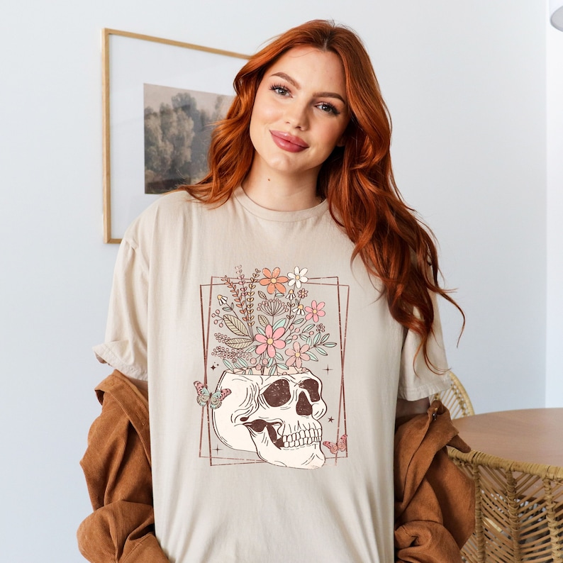 May include: A beige t-shirt with a graphic of a skull with flowers growing out of the top. The skull is outlined in brown and the flowers are pink, orange, and white.