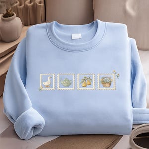 A light blue sweater with a row of four small illustrations on it, including a duck, teapot, pears, and a basket.