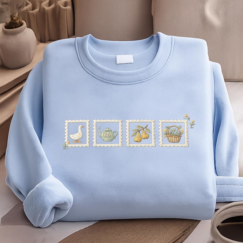 A light blue sweater with a row of four small illustrations on it, including a duck, teapot, pears, and a basket.