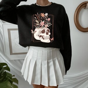 May include: Black cropped sweatshirt with a graphic of a skull with flowers growing out of the top. The skull is surrounded by a gold frame and there are butterflies on either side of the skull.