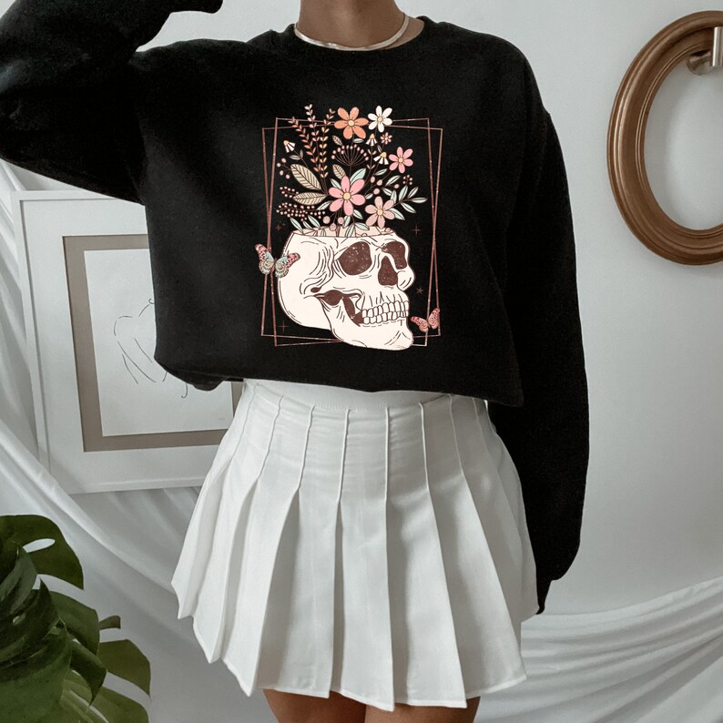May include: Black cropped sweatshirt with a graphic of a skull with flowers growing out of the top. The skull is surrounded by a gold frame and there are butterflies on either side of the skull.