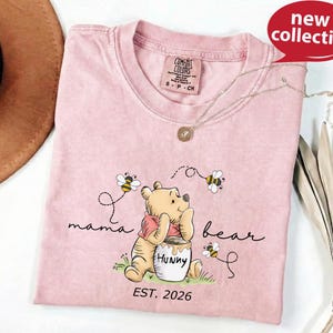 A pink t-shirt with a Winnie the Pooh design and the text "mama bear" printed on it, along with a necklace and a brown hat.