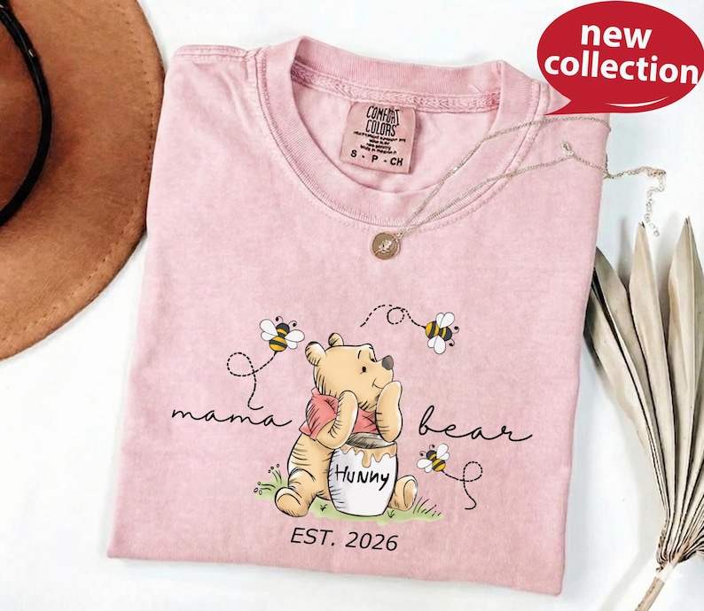 A pink t-shirt with a Winnie the Pooh design and the text "mama bear" printed on it, along with a necklace and a brown hat.