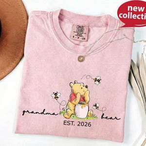A pink t-shirt with a Winnie the Pooh graphic and the text "Grandma Bear" printed on it, along with the text "EST. 2026" below the image.