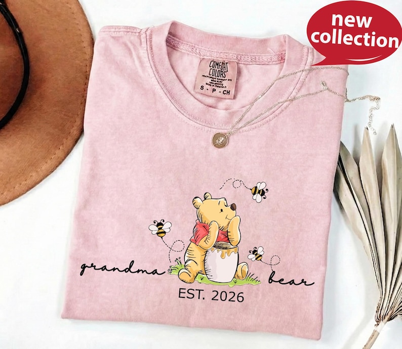 A pink t-shirt with a Winnie the Pooh graphic and the text "Grandma Bear" printed on it, along with the text "EST. 2026" below the image.