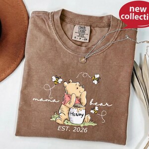 A brown t-shirt with a Winnie the Pooh-themed design, including the character Huckleberry Bear, is displayed on a white surface.