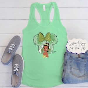 May include: A mint green vest top with a graphic of Princess Tiana inside a Mickey Mouse head outline with a green bow. The text "Tiana" is below the princess. Grey trainers and denim jeans are also in the image.