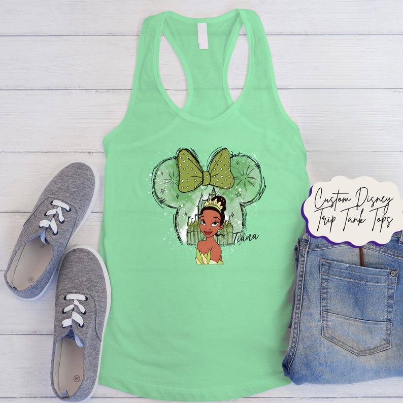 May include: A mint green vest top with a graphic of Princess Tiana inside a Mickey Mouse head outline with a green bow. The text "Tiana" is below the princess. Grey trainers and denim jeans are also in the image.