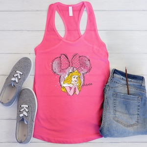 May include: A vibrant pink vest top with a Princess Aurora graphic. The design incorporates a Minnie Mouse-style bow and ears, with the name "Aurora" in script. Grey trainers and blue jeans are also in the image.